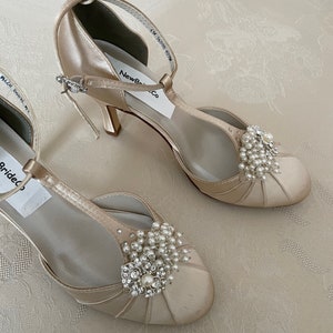 May include: A pair of ivory satin bridal shoes with a t-strap design and a small heel. The shoes feature a pearl and rhinestone embellishment on the toe.