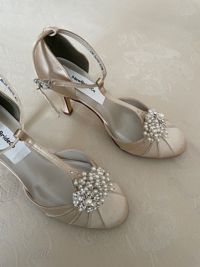 May include: A pair of ivory satin bridal shoes with a t-strap design and a small heel. The shoes feature a pearl and rhinestone embellishment on the toe.