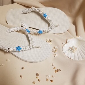 May include: White flip flops decorated with white crocheted straps, blue starfish, and white pearls. The flip flops are on a beige surface with small white and brown seashells scattered around.