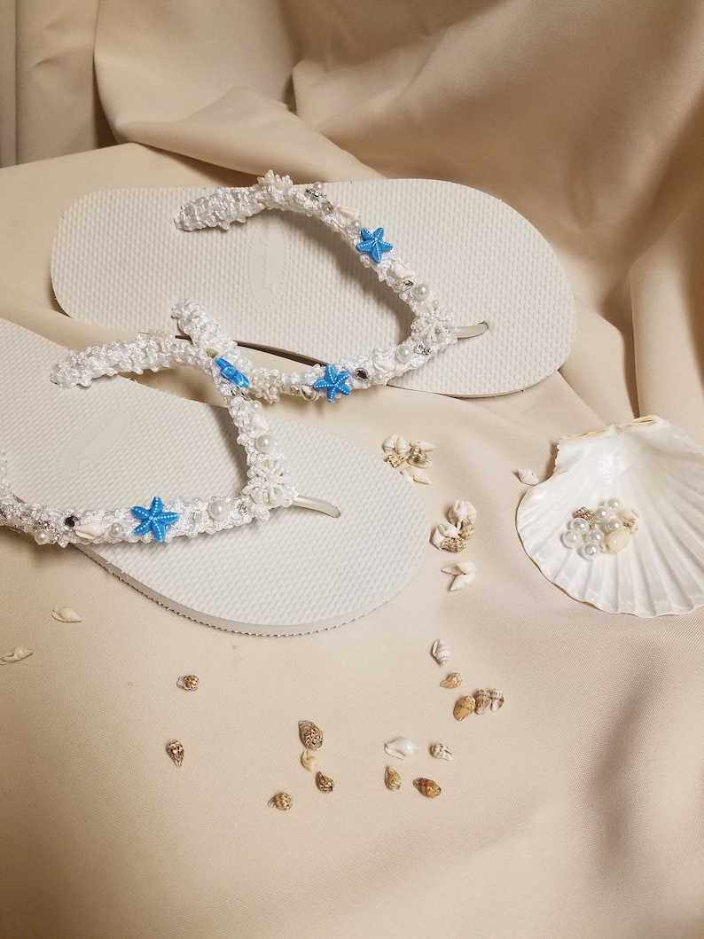 May include: White flip flops decorated with white crocheted straps, blue starfish, and white pearls. The flip flops are on a beige surface with small white and brown seashells scattered around.