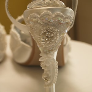May include: A white satin high-heeled shoe featuring an ornate heel embellished with intricate white lace, iridescent sequins, small round pearls, and clear crystals. The heel design includes a floral-shaped applique and a delicate beaded trim.