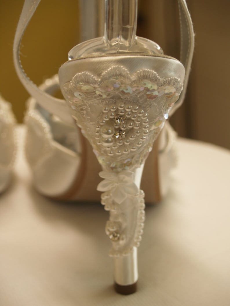 May include: A white satin high-heeled shoe featuring an ornate heel embellished with intricate white lace, iridescent sequins, small round pearls, and clear crystals. The heel design includes a floral-shaped applique and a delicate beaded trim.