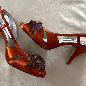 Puede incluir: Un par de sandalias de tacón de satén naranja con adornos florales. Las sandalias tienen la punta abierta y una correa de tobillo.