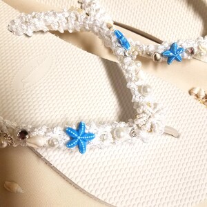 May include: White flip flops with a white crocheted strap decorated with blue starfish, white pearls, and seashells.