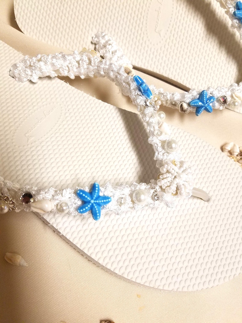 May include: White flip flops with a white crocheted strap decorated with blue starfish, white pearls, and seashells.