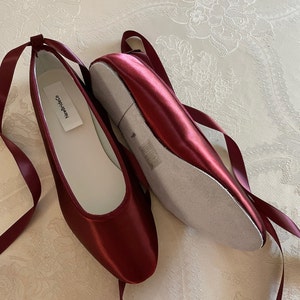 Peut inclure: Une paire de ballerines en satin bordeaux avec des rubans assortis. Les chaussures ont un bout arrondi et une semelle intérieure blanche. L'étiquette à l'intérieur d'une chaussure indique "NewBridco.co".