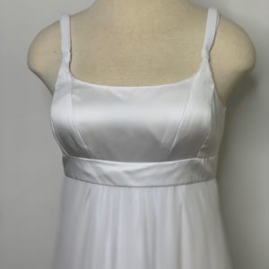 Puede incluir: Un vestido de satén blanco con escote cuadrado y tirantes finos. El corpiño tiene un diseño entallado con una cintura definida. La falda está hecha de una tela fluida y transparente, creando un aspecto suave y elegante. El vestido se exhibe en un maniquí.