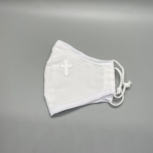 May include: White face mask with a small, embroidered cross on the front. The mask has adjustable white cords and is made of a soft, breathable fabric. The mask is designed to cover the nose and mouth.