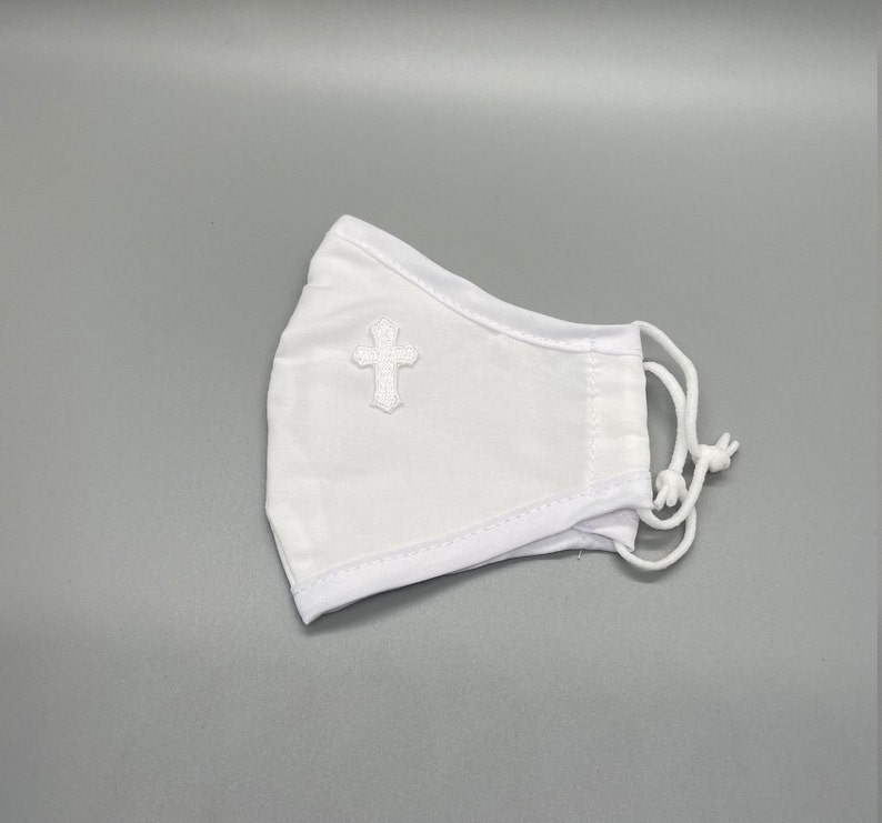 May include: White face mask with a small, embroidered cross on the front. The mask has adjustable white cords and is made of a soft, breathable fabric. The mask is designed to cover the nose and mouth.