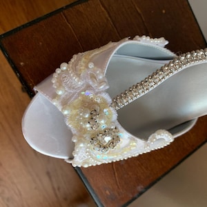 May include: A white open-toe shoe featuring an embellished strap adorned with clear rhinestones, white pearls, and intricate lace floral appliqu&eacute;s. The design includes iridescent sequins and a central cluster of sparkling crystals on the upper.