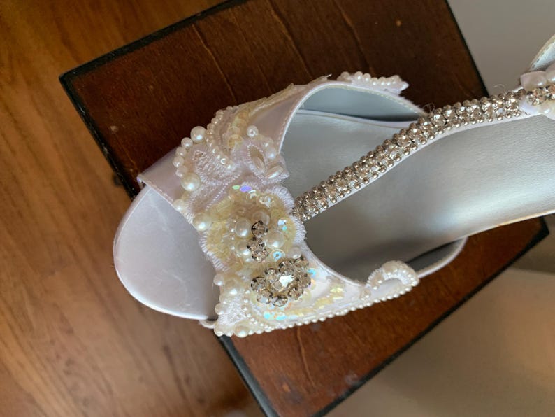 May include: A white open-toe shoe featuring an embellished strap adorned with clear rhinestones, white pearls, and intricate lace floral appliqu&eacute;s. The design includes iridescent sequins and a central cluster of sparkling crystals on the upper.