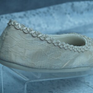 Puede incluir: Una bailarina de encaje blanco con un delicado ribete plateado. El zapato est&aacute; en un expositor de acr&iacute;lico transparente.