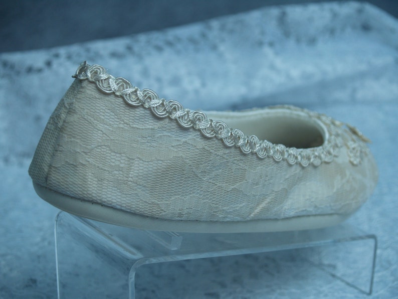 Puede incluir: Una bailarina de encaje blanco con un delicado ribete plateado. El zapato est&aacute; en un expositor de acr&iacute;lico transparente.