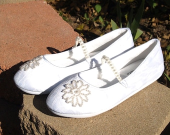 Puede incluir: Un par de zapatos de tela blanca con adornos de perlas. Un zapato tiene un diseño de flor de perlas, y el otro tiene una correa de perlas.