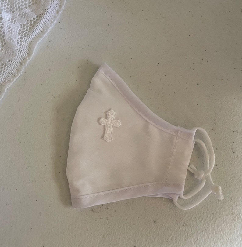 May include: White face mask with a white embroidered cross on the front. The mask has white trim and adjustable white ear loops. A piece of white lace is visible in the background.
