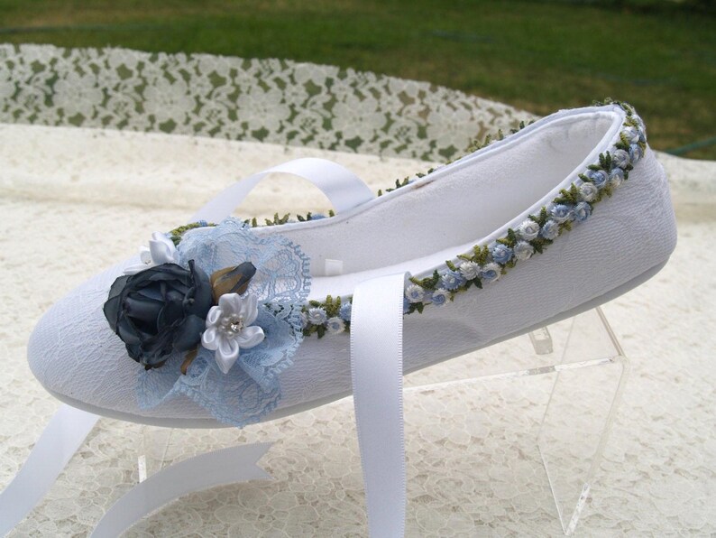 May include: White satin slipper with blue and green floral embroidery. The slipper has a white satin ribbon and a blue and white floral embellishment.
