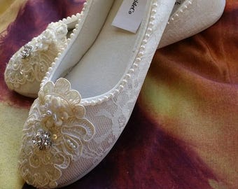 May include: A pair of white lace ballet flats featuring a delicate pearl trim along the collar and a floral lace applique on the toe adorned with clear crystals and small pearls. A white tag labeled NewBrideCo is visible inside the shoe.