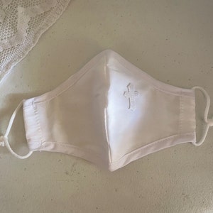 May include: White face mask with an embroidered cross. The mask has adjustable ear loops and is made of a soft, white fabric. A piece of white lace is visible in the upper left corner. The mask is designed for personal use.