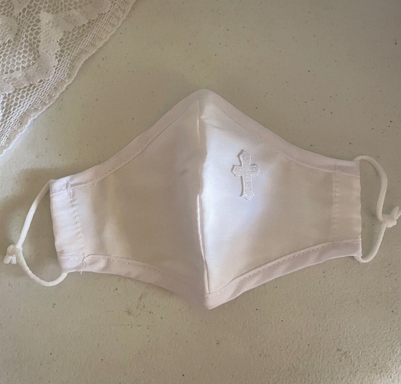 May include: White face mask with an embroidered cross. The mask has adjustable ear loops and is made of a soft, white fabric. A piece of white lace is visible in the upper left corner. The mask is designed for personal use.