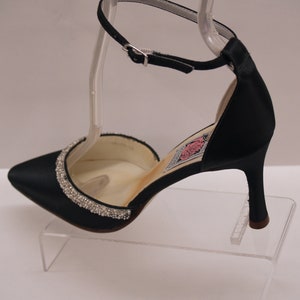 May include: Black satin heeled shoe with a rhinestone trim around the toe and an ankle strap. The shoe is on a clear display stand.