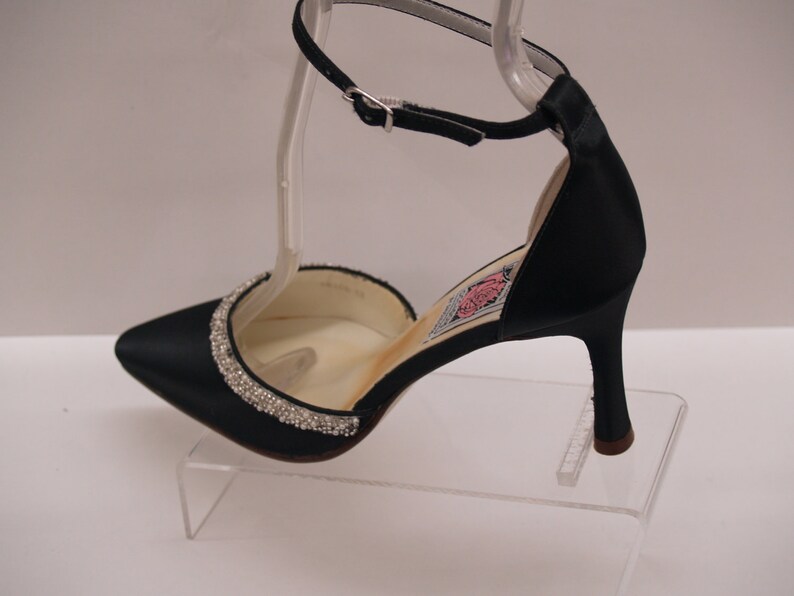 May include: Black satin heeled shoe with a rhinestone trim around the toe and an ankle strap. The shoe is on a clear display stand.