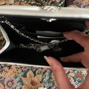 May include: A white clutch bag with a silver metal chain strap. The interior is lined in black fabric and features a small slip pocket. A white label with black text is visible inside the main compartment, along with a folded white paper insert.