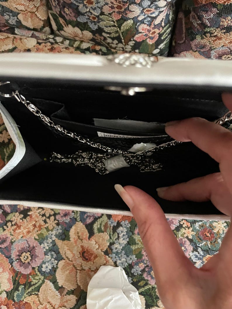 May include: A white clutch bag with a silver metal chain strap. The interior is lined in black fabric and features a small slip pocket. A white label with black text is visible inside the main compartment, along with a folded white paper insert.