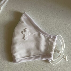 May include: White face mask with a small, embroidered cross on the front. The mask has adjustable white cords and a soft, satin-like texture. The mask is designed to cover the nose and mouth.