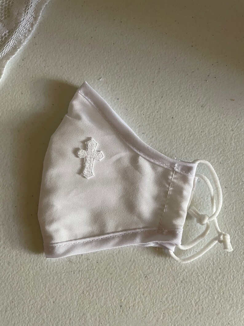 May include: White face mask with a small, embroidered cross on the front. The mask has adjustable white cords and a soft, satin-like texture. The mask is designed to cover the nose and mouth.
