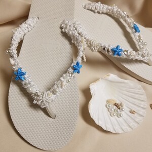 May include: White flip flops with a white crocheted design featuring blue starfish and white beads. The flip flops are on a beige surface with a white seashell.