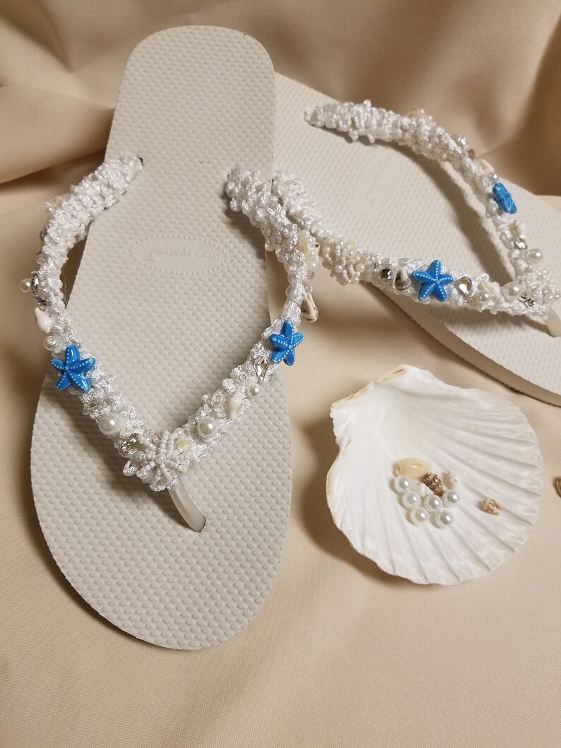 May include: White flip flops with a white crocheted design featuring blue starfish and white beads. The flip flops are on a beige surface with a white seashell.