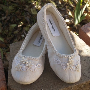 Puede incluir: Un par de zapatos de novia de sat&eacute;n blanco con un delicado borde de encaje y adornos florales. Los zapatos est&aacute;n etiquetados "NewBrideCo."