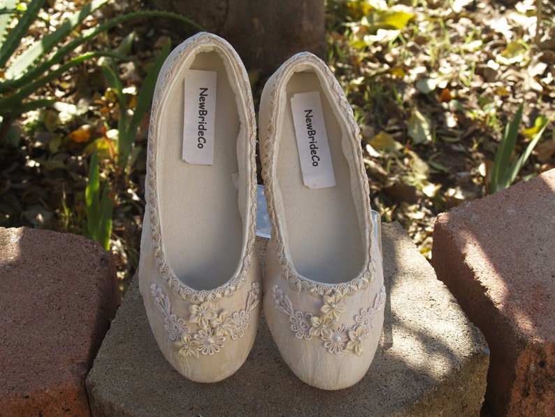 Puede incluir: Un par de zapatos de novia de sat&eacute;n color marfil con delicado bordado floral y una etiqueta NewBrideCo. Los zapatos est&aacute;n sobre una superficie de piedra.