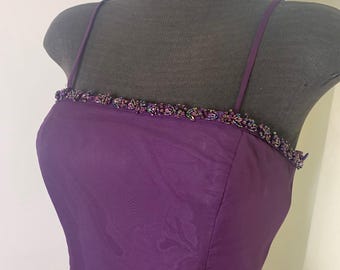 May include: A sleeveless, floor-length purple dress with thin straps. The neckline is embellished with a beaded trim. The dress is displayed on a mannequin, showcasing its elegant design and flowing fabric.