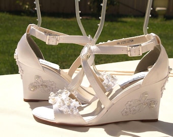 Può includere: Un paio di sandali con zeppa in raso bianco con cinturino alla caviglia con fibbia color argento, punta aperta e applicazioni decorative in pizzo floreale adornate con piccole perle bianche e cristalli trasparenti sul tallone.
