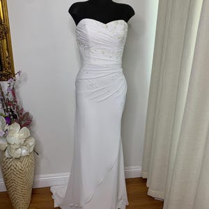 May include: A white strapless wedding dress on a black mannequin. The bodice features delicate embroidery and ruching, while the skirt flows to the floor. The dress is displayed in a well-lit room with a vase of flowers and a mirror in the background.