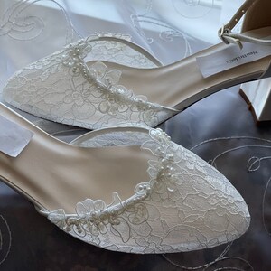 May include: Ivory lace bridal shoes with floral and pearl accents. The shoes have a pointed toe and an ankle strap with a buckle. The shoes are new and have a tag that says "NewBrideCo".
