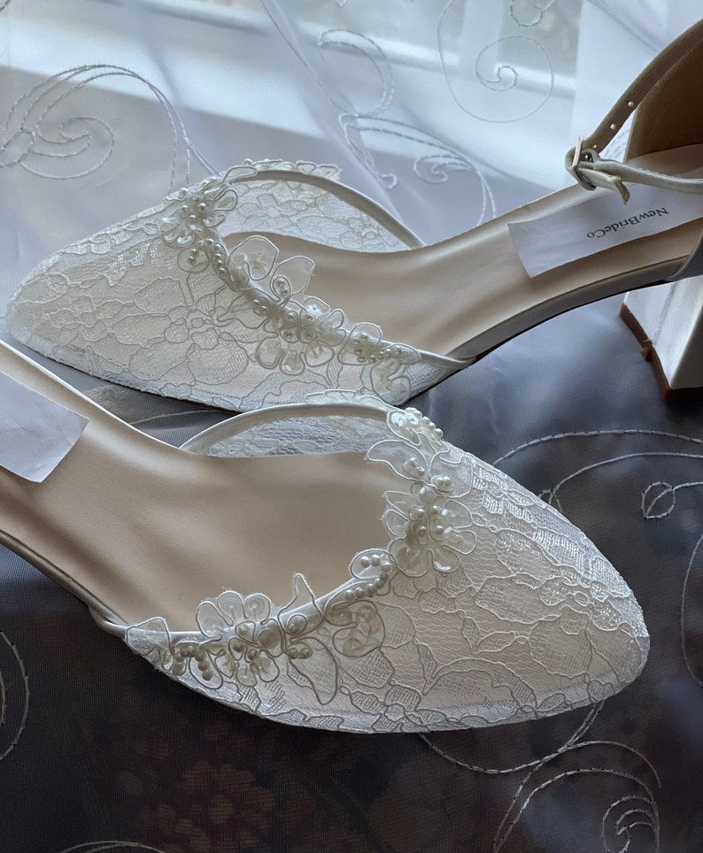 May include: Ivory lace bridal shoes with floral and pearl accents. The shoes have a pointed toe and an ankle strap with a buckle. The shoes are new and have a tag that says "NewBrideCo".