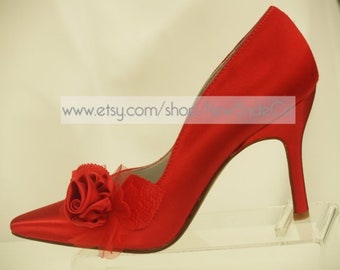 Puede incluir: Zapatos de tacón alto de satén rojo con un gran adorno de rosa roja en la punta. Los zapatos están sobre un expositor de acrílico transparente.
