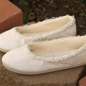 May include: A pair of white satin slippers with a delicate beaded trim. The slippers have a rounded toe and a low heel.