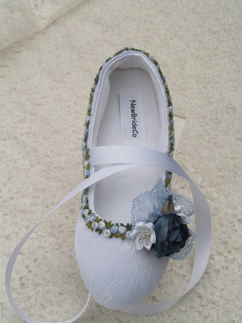 May include: White satin bridal shoes with blue and green embroidery, a blue satin flower, and a white satin ribbon. The shoes have the text "NewBrideCo" printed on the inside.