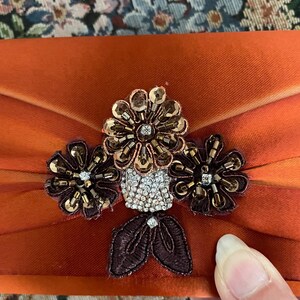 May include: A burnt orange satin clutch bag featuring a central floral embellishment. The ornament consists of three dark brown beaded flowers with metallic sequins, a cluster of clear rhinestones, and two dark brown embroidered leaves.