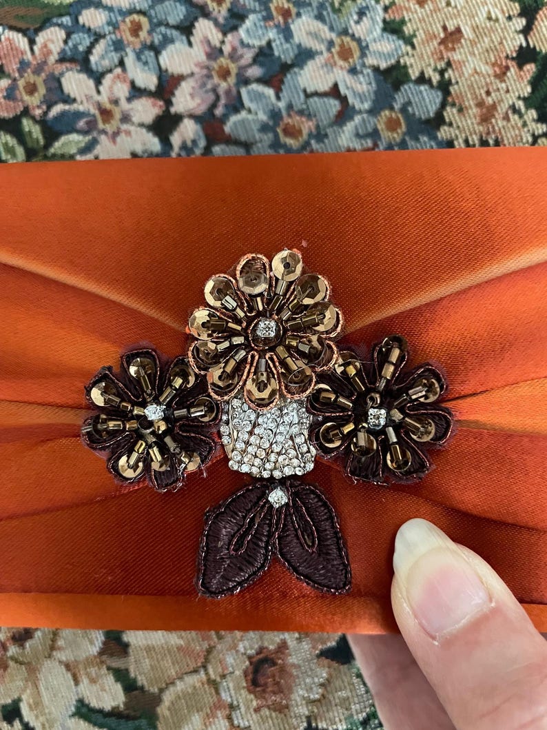 May include: A burnt orange satin clutch bag featuring a central floral embellishment. The ornament consists of three dark brown beaded flowers with metallic sequins, a cluster of clear rhinestones, and two dark brown embroidered leaves.
