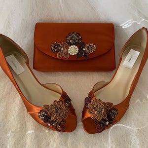 May include: A pair of burnt orange satin peep-toe heels and a matching rectangular clutch bag. Both items feature floral appliqu&eacute;s with brown, bronze, and clear crystal beadwork. Each shoe and the clutch bag include a white NewBrideCo brand label.