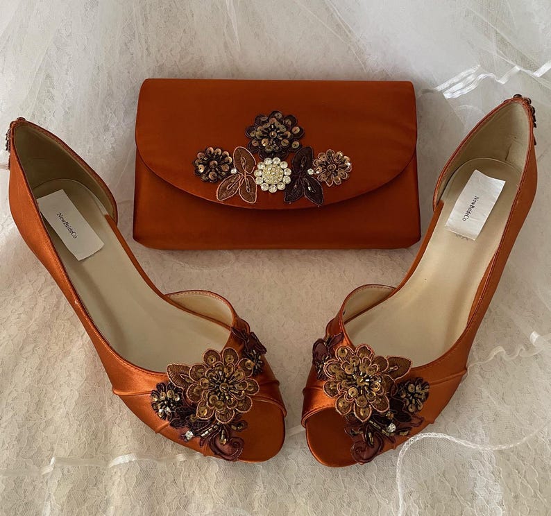May include: A pair of burnt orange satin peep-toe heels and a matching rectangular clutch bag. Both items feature floral appliqu&eacute;s with brown, bronze, and clear crystal beadwork. Each shoe and the clutch bag include a white NewBrideCo brand label.