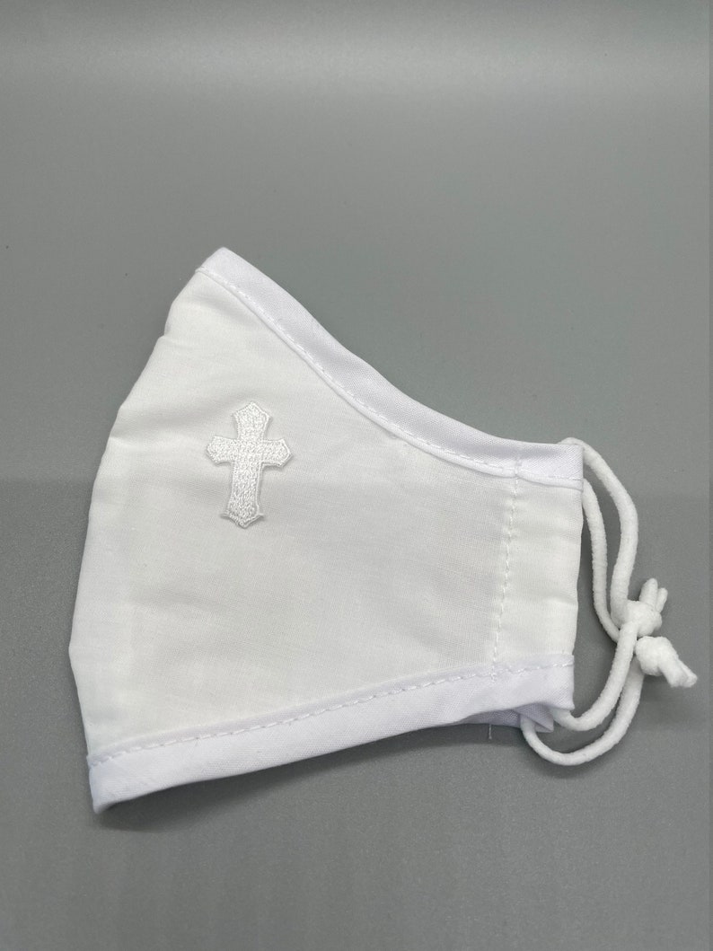 May include: White face mask with a white embroidered cross on the front. The mask has white trim and adjustable white cords. The mask is made of a soft, breathable fabric.