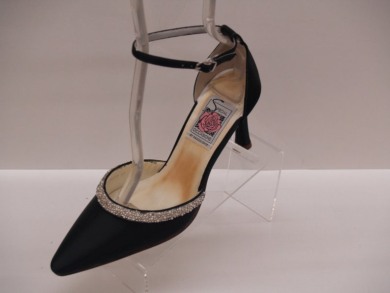 May include: Black satin heeled shoe with a rhinestone trim around the toe and an ankle strap. The shoe has a label that reads "Special Occasions" with a pink rose.