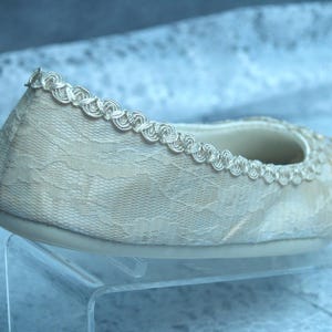Op de afbeelding: Een witte platte schoen van kant met een delicate kanten rand rondom. De schoen staat op een witte displaystandaard.