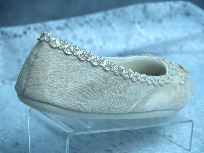Op de afbeelding: Een witte platte schoen van kant met een delicate kanten rand rondom. De schoen staat op een witte displaystandaard.