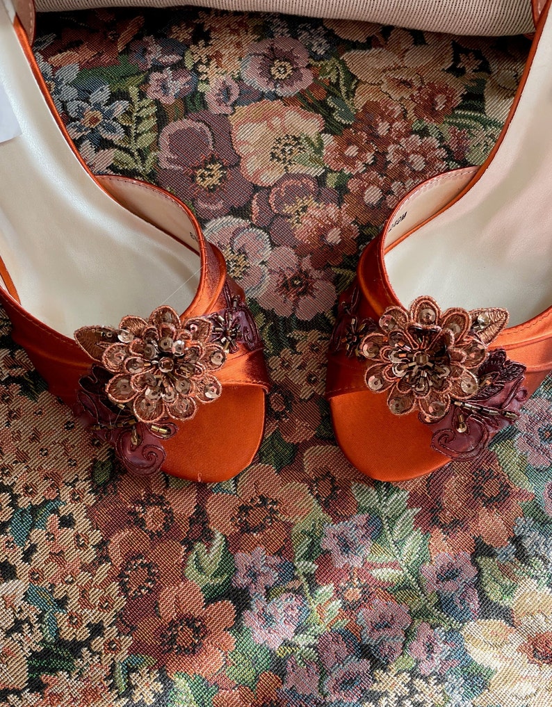May include: A pair of burnt orange satin open-toe shoes featuring a decorative floral appliqu&eacute; on the front. Each flower is crafted from layered fabric petals, dark brown embroidery, and small metallic sequins that catch the light.
